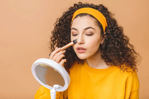 Perfect view. Morning routine. Joyful young curly woman is sapplying foundation cream on her skin. She is looking at mirror in her hand. Isolated on beige background
