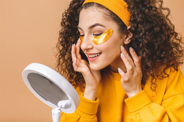 Image of young pure beautiful curly woman isolated over beige background take care of her skin with under eye patches.
