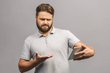 Memnun olmayan sakallı adam internette araştırma yapıyor, tabletten haberler okuyor, tatminsizlikten kaşlarını çatıyor, resmi tişört giyiyor, gri duvarda izole edilmiş. Olumsuz duygular