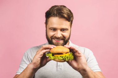 Hamburger tutan genç bir adam. Sakallı Gyu fast food yiyor. Burger yararlı bir yemek değildir. Çok aç bir adam. Diyet konsepti. Pembe arka planda izole edilmiş.