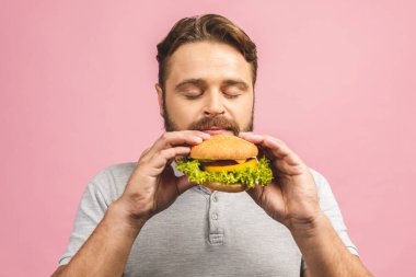 Hamburger tutan genç bir adam. Sakallı Gyu fast food yiyor. Burger yararlı bir yemek değildir. Çok aç bir adam. Diyet konsepti. Pembe arka planda izole edilmiş.