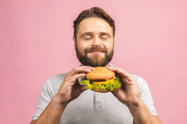 Hamburger tutan genç bir adam. Sakallı Gyu fast food yiyor. Burger yararlı bir yemek değildir. Çok aç bir adam. Diyet konsepti. Pembe arka planda izole edilmiş.