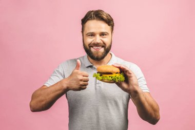 Hamburger tutan genç bir adam. Sakallı Gyu fast food yiyor. Burger yararlı bir yemek değildir. Çok aç bir adam. Diyet konsepti. Pembe arka planda izole edilmiş. Başparmaklar yukarı.. 