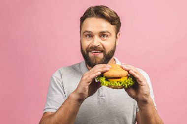 Hamburger tutan genç bir adam. Sakallı Gyu fast food yiyor. Burger yararlı bir yemek değildir. Çok aç bir adam. Diyet konsepti. Pembe arka planda izole edilmiş. 
