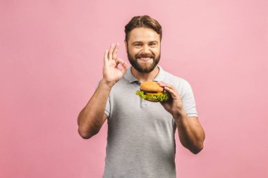 Hamburger tutan genç bir adam. Sakallı Gyu fast food yiyor. Burger yararlı bir yemek değildir. Çok aç bir adam. Diyet konsepti. Pembe arka planda izole edilmiş. Tamam imzala.