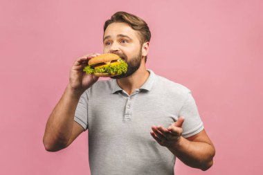 Hamburger tutan genç bir adam. Sakallı Gyu fast food yiyor. Burger yararlı bir yemek değildir. Çok aç bir adam. Diyet konsepti. Pembe arka planda izole edilmiş.