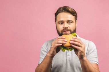 Hamburger tutan genç bir adam. Sakallı Gyu fast food yiyor. Burger yararlı bir yemek değildir. Çok aç bir adam. Diyet konsepti. Pembe arka planda izole edilmiş.