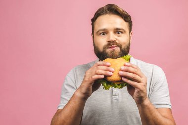 Hamburger tutan genç bir adam. Sakallı Gyu fast food yiyor. Burger yararlı bir yemek değildir. Çok aç bir adam. Diyet konsepti. Pembe arka planda izole edilmiş.