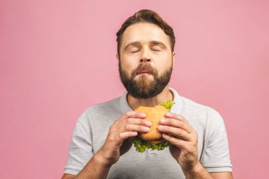 Hamburger tutan genç bir adam. Sakallı Gyu fast food yiyor. Burger yararlı bir yemek değildir. Çok aç bir adam. Diyet konsepti. Pembe arka planda izole edilmiş.