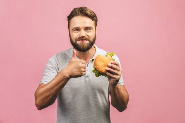 Hamburger tutan genç bir adam. Sakallı Gyu fast food yiyor. Burger yararlı bir yemek değildir. Çok aç bir adam. Diyet konsepti. Pembe arka planda izole edilmiş. Başparmaklar yukarı..
