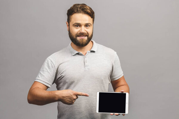 Cheerful bearded young man in casual pointing on blank screen tablet isolated over grey background.