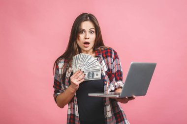 Elinde para banknotları tutan neşeli genç bir kadının portresi ve pembe arka planda izole edilmiş kutlamayı. Laptop kullanılıyor.