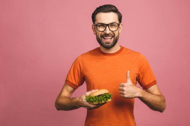 Hamburger tutan genç bir adam. Öğrenci fast food yiyor. Burger yararlı bir yemek değildir. Çok aç bir adam. Diyet konsepti. Pembe arka planda izole edilmiş. Başparmaklar yukarı.. 