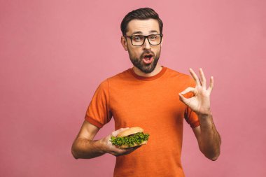 Hamburger tutan genç bir adam. Öğrenci fast food yiyor. Burger yararlı bir yemek değildir. Çok aç bir adam. Diyet konsepti. Pembe arka planda izole edilmiş. Tamam imzala. 