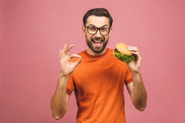 Hamburger tutan genç bir adam. Öğrenci fast food yiyor. Burger yararlı bir yemek değildir. Çok aç bir adam. Diyet konsepti. Pembe arka planda izole edilmiş. Tamam imzala. 