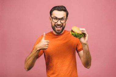 Hamburger tutan genç bir adam. Öğrenci fast food yiyor. Burger yararlı bir yemek değildir. Çok aç bir adam. Diyet konsepti. Pembe arka planda izole edilmiş. Başparmaklar yukarı.. 