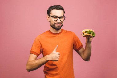 Hamburger tutan genç bir adam. Öğrenci fast food yiyor. Burger yararlı bir yemek değildir. Çok aç bir adam. Diyet konsepti. Pembe arka planda izole edilmiş. Başparmaklar yukarı.. 