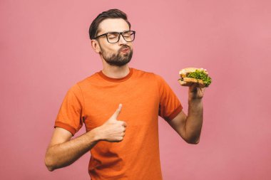 Hamburger tutan genç bir adam. Öğrenci fast food yiyor. Burger yararlı bir yemek değildir. Çok aç bir adam. Diyet konsepti. Pembe arka planda izole edilmiş. Başparmaklar yukarı.. 