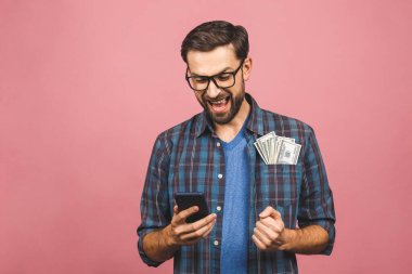 Mutlu galip! Kareli gömlekli genç adam cebinde dolar banknotları tutuyor ve sürpriz bir şekilde pembe arka planda izole edilmiş. Telefon kullanılıyor. 