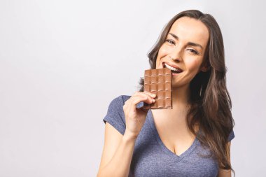 Çikolata yiyip gülümseyen genç ve mutlu bir bayan. Tatlı çikolatanın tadına bakan bir kız. Doğal makyajlı genç bir kadın beyaz arka planda izole edilmiş çikolata yiyip eğleniyor.