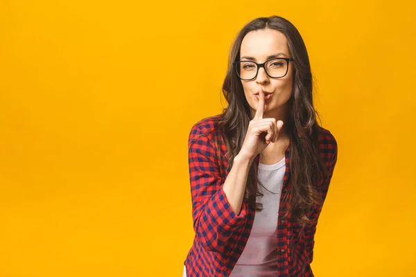 It\'s a secret! Shut your mouth. Finger on lips. Portrait of pretty cheerful female making shush or shh gesture with index finger over mouth, looking confident at camera over yellow background.