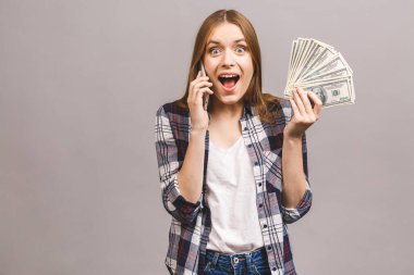 Mutlu, heyecanlı bir kadının portresi. Gri arka planda izole edilmiş bir sürü para banknotunu tutarken cep telefonuyla konuşuyor..