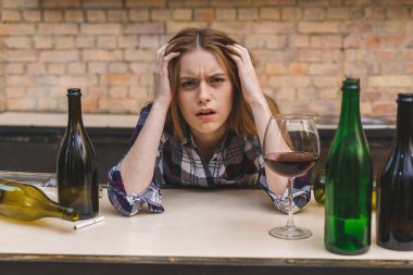 Genç üzgün ve sarhoş bir kadın mutfak koltuğunda oturmuş kırmızı şarap içiyor, tamamen sarhoş görünüyor yalnız görünüyor ve alkolizm ve alkol bağımlılığından muzdarip..