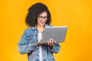 Genç Afro-Amerikan siyah, kıvırcık saçlı, dizüstü bilgisayar kullanan ve sarı arka planda sırıtan havalı bayan.. 