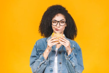Sarı arka planda izole edilmiş hamburger yiyen Afro-Amerikalı genç kadın..