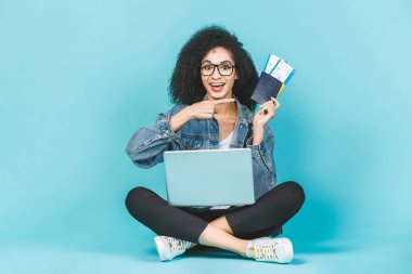 Yerde dizüstü bilgisayarı ve mavi arka planda izole edilmiş uçak biletleriyle oturan oldukça genç, mutlu bir siyah Afrikalı Amerikalı kadın. İşaret parmağı.
