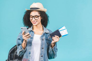 Stüdyoda mavi arka planda izole edilmiş mutlu Afrikalı Amerikalı kız. Seyahat konsepti. Pasaport, bilet. Telefon kullanılıyor.