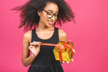 Mutlu Afro-Amerikan kıvırcık bayanı pembe arka planda izole edilmiş hediyesini tutarken gülerken.