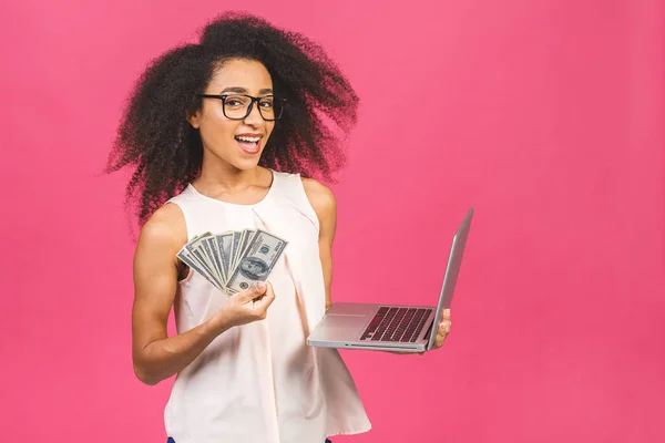 Happy winner. Portrait of african american successful woman 20s with afro hairstyle holding lots of money dollar banknotes and laptop isolated over pink background.