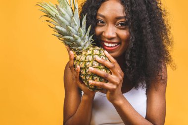 Ananas egzotik orta saha oyuncusu diyette. Afro-Amerikan siyah esmer tenli neşeli kız, sarı arka planda izole edilmiş ananası elinde tutuyor..