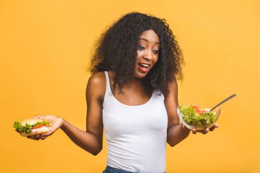 Sağlıklı gıda konsepti. Sarı arka planda izole edilmiş siyah salata ve burger yiyen güzel Afro-Amerikalı kadının yakın çekimi. 