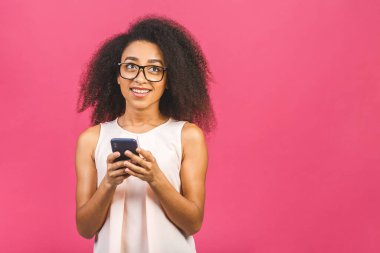 Mutlu, genç, Afro-Amerikan bir kadın. Sıradan giyinmiş, pembe arka planda tek başına duruyor. Elinde cep telefonu var..