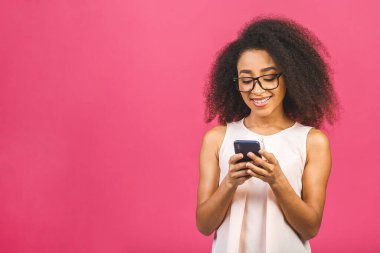 Mutlu, genç, Afro-Amerikan bir kadın. Sıradan giyinmiş, pembe arka planda tek başına duruyor. Elinde cep telefonu var..