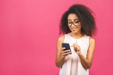 Mutlu, genç, Afro-Amerikan bir kadın. Sıradan giyinmiş, pembe arka planda tek başına duruyor. Elinde cep telefonu var..