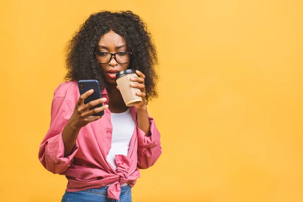 Happy young african american woman casually dressed standing isolated over yellow background, talking on mobile phone, holding takeaway cup of coffee.
