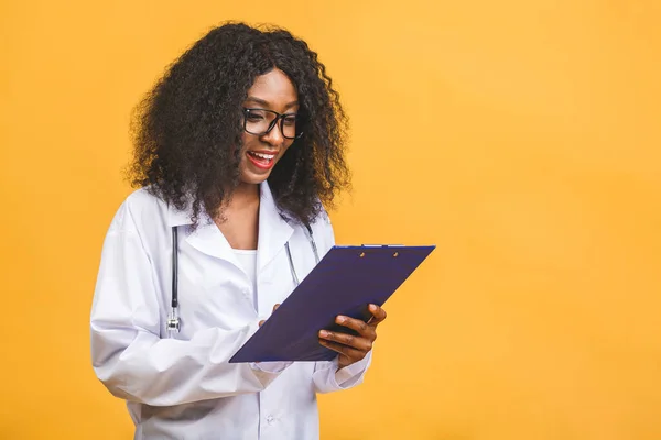 Medicine, profession and healthcare concept - smiling african american female doctor in white coat with clipboard and stethoscope isolated over yellow background.