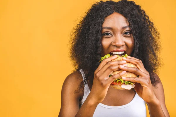 Sarı arka planda izole edilmiş hamburger yiyen Afro-Amerikalı güzel genç kadın..