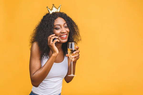 Happy birthday! Portrait of happy African-American black woman with glass of champagne and golden crown isolated over yellow background. Using phone.