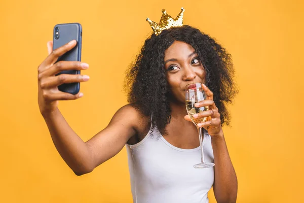 Happy birthday! Portrait of happy African-American black woman with glass of champagne and golden crown isolated over yellow background. Using phone.