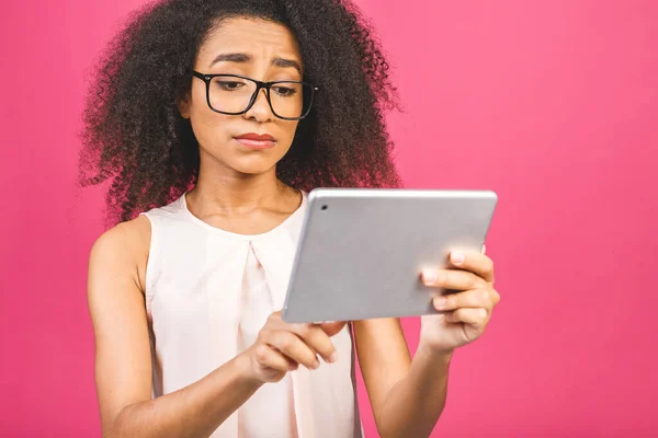 Isolated sad young american student girl with curly african hair holding digital tablet standing over isolated pink background with copy space for text, logo or advertising.