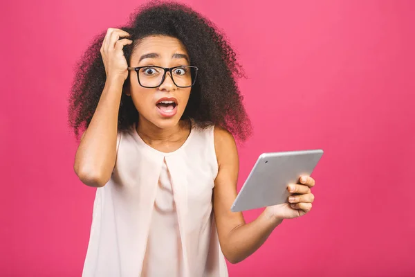 Shocked american student girl with curly african hair holding digital tablet over isolated pink background with copy space for text, logo or advertising.