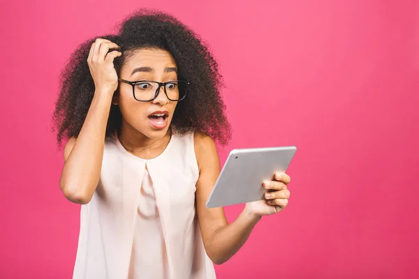 Shocked american student girl with curly african hair holding digital tablet over isolated pink background with copy space for text, logo or advertising.