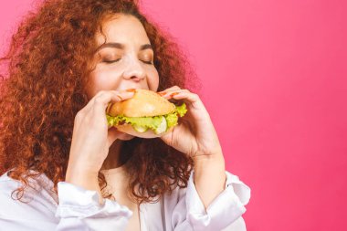 Abur cubur konsepti. Çekici kıvırcık saçlı beyaz kadın pembe arka planda izole edilmiş büyük bir hamburger tutuyor ve yiyor.. 