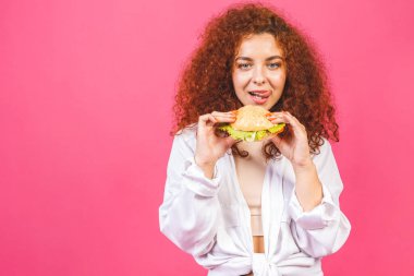 Abur cubur konsepti. Çekici kıvırcık saçlı beyaz kadın pembe arka planda izole edilmiş büyük bir hamburger tutuyor ve yiyor.. 