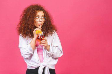 Renkli pembe arka planda izole edilmiş bir bardak kokteyl içeceği stüdyosuyla yaz kıyafetleri içinde mutlu, güzel kıvırcık kadın..