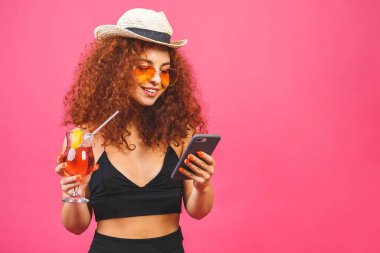 Akıllı telefon kullanan ve pembe arka planda izole edilmiş kokteyl tutan güneş gözlüklü mutlu kadın portresi. 